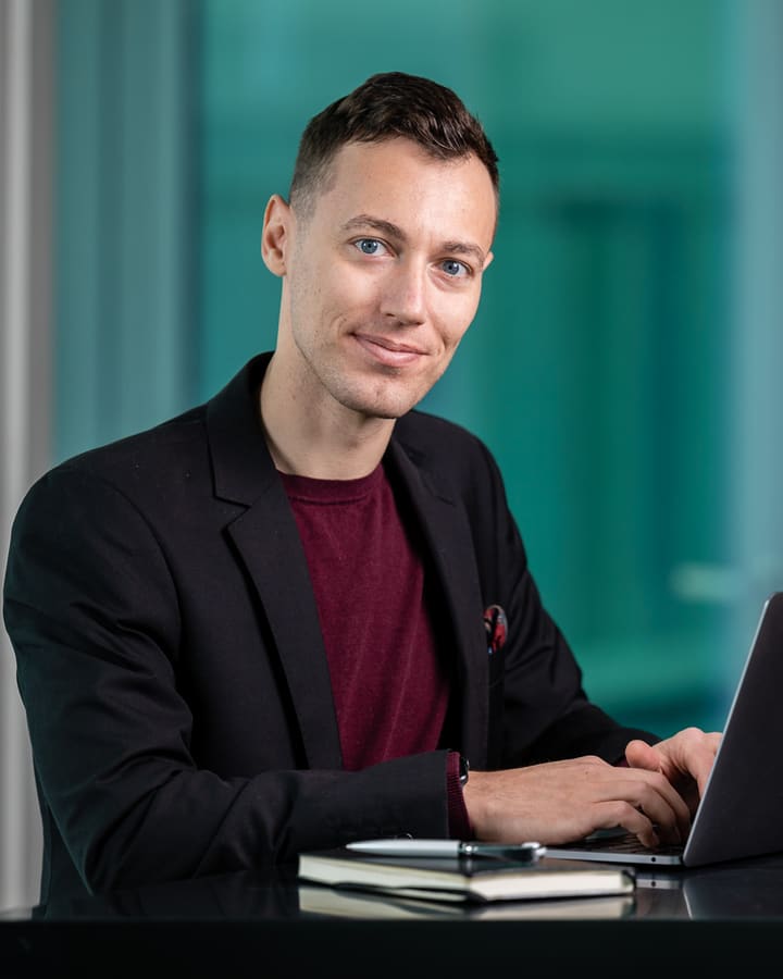 Adrian Ley am Schreibtisch vor großen Fenstern, lächelt in die Kamera